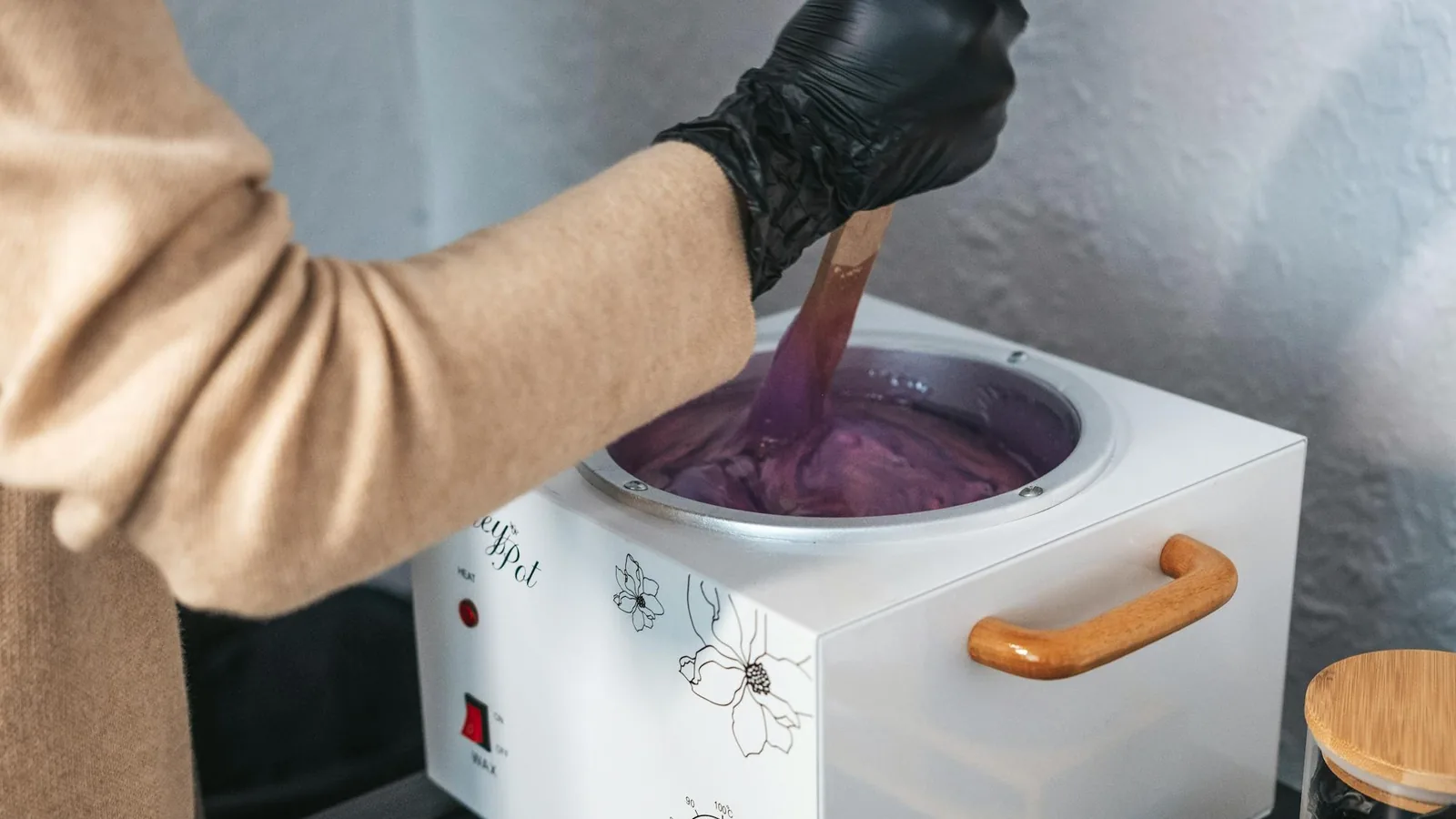 Esthetician stirring warm wax in a treatment pot — soft wax adheres to both hair and skin, unlike sugar paste
